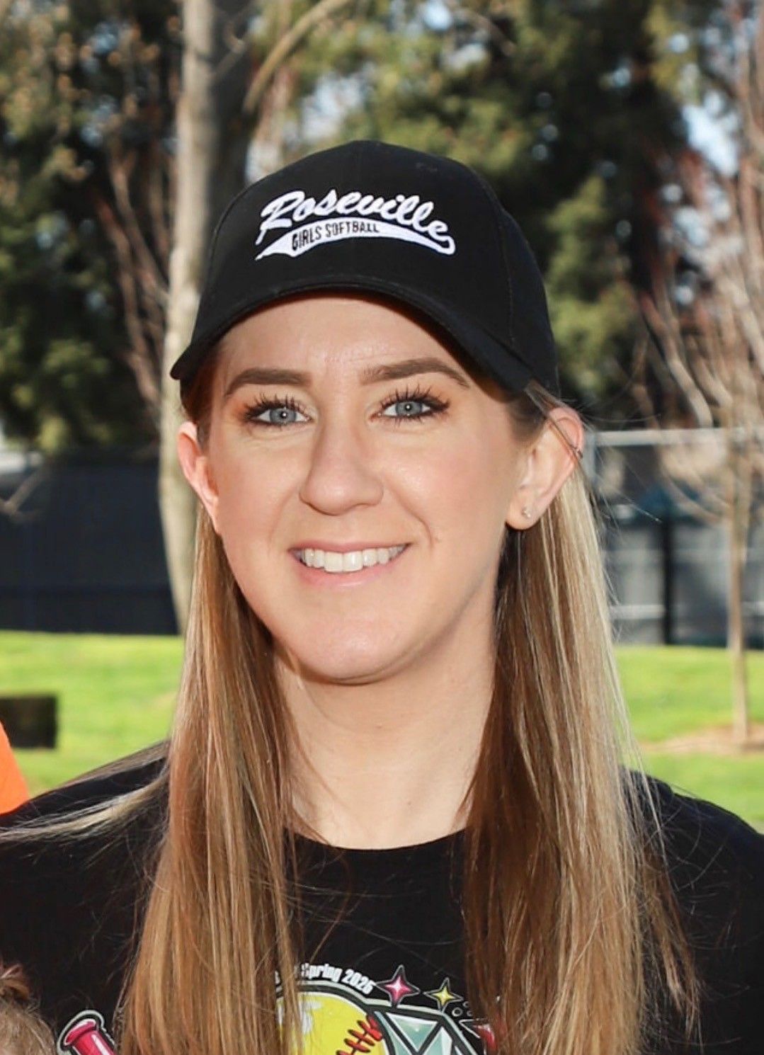 Smiling woman in a black cap and shirt outdoors, with trees and a fence in the background