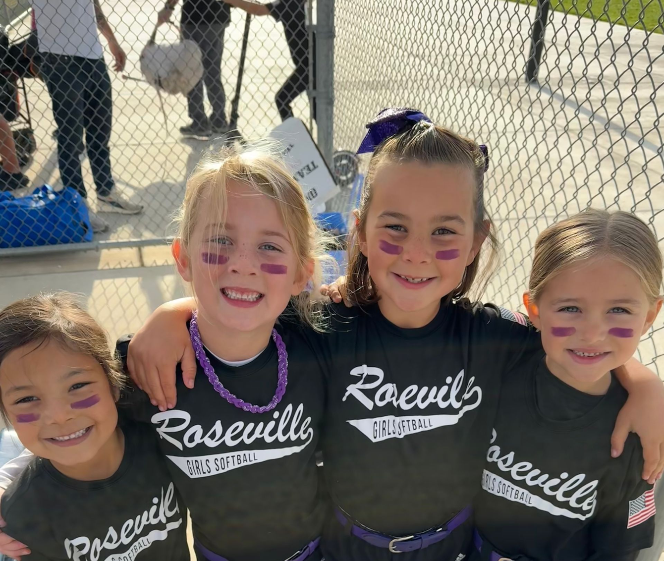 Four smiling kids in black Roseville shirts with face paint, standing by a chain-link fence at a sports field.