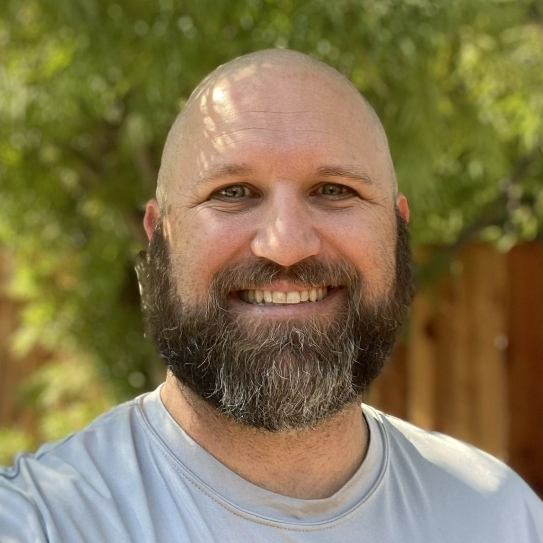 Smiling bald man with a beard outdoors, wearing a light gray shirt, with trees in the background