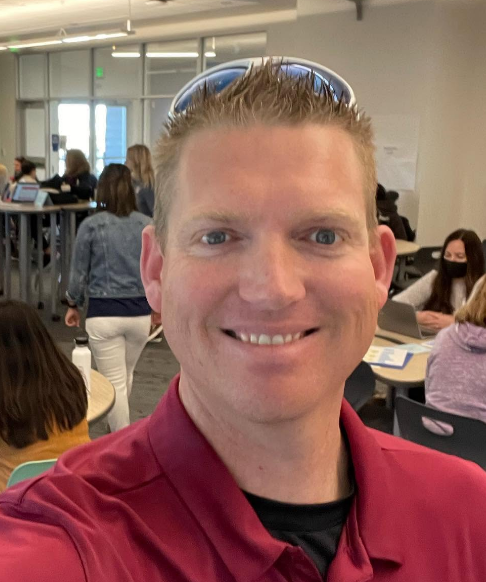 Smiling person in red polo taking a selfie in a busy indoor classroom or office.