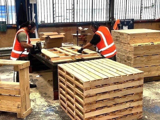 Large warehouse interior with stacked wooden pallets and crates under bright overhead lights