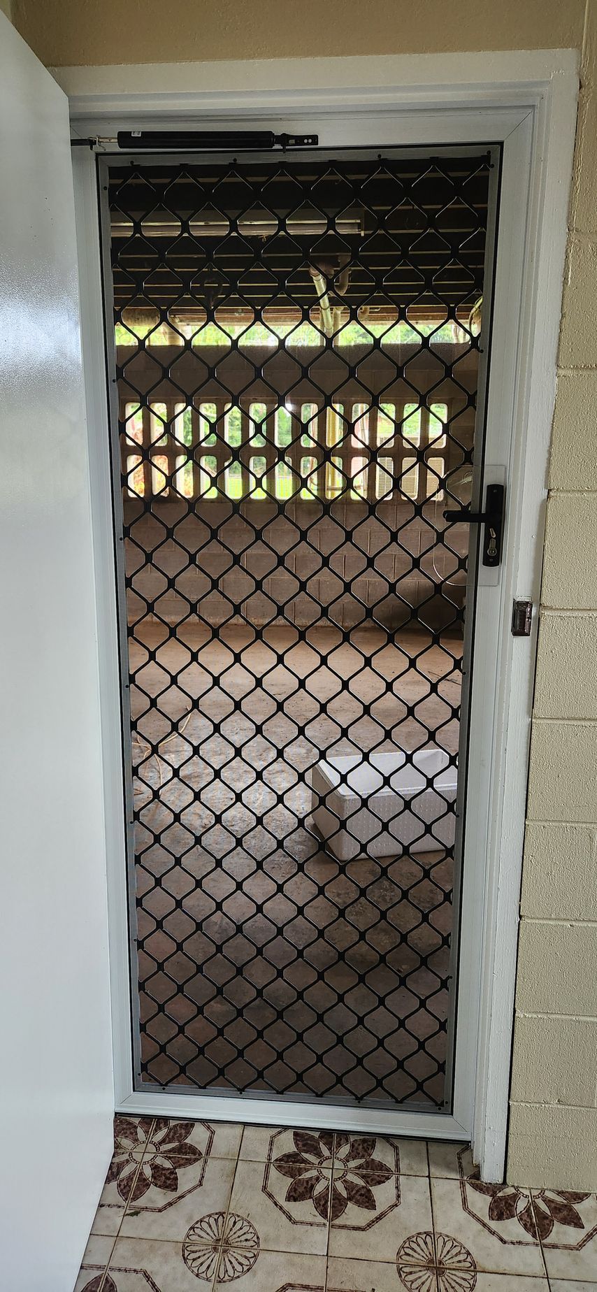 A metal screen door with diamond-shaped mesh, leading to a blurred outdoor view, set in a doorway.- Advancetech Glass & Glazing in Wulguru, QLD