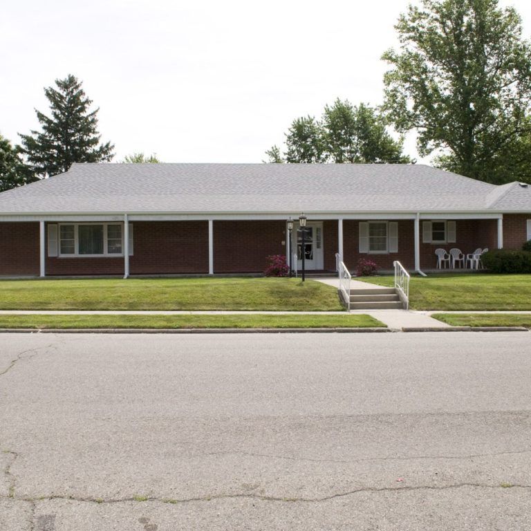 Single-story red brick building with white trim, front steps, and a lawn beside the street