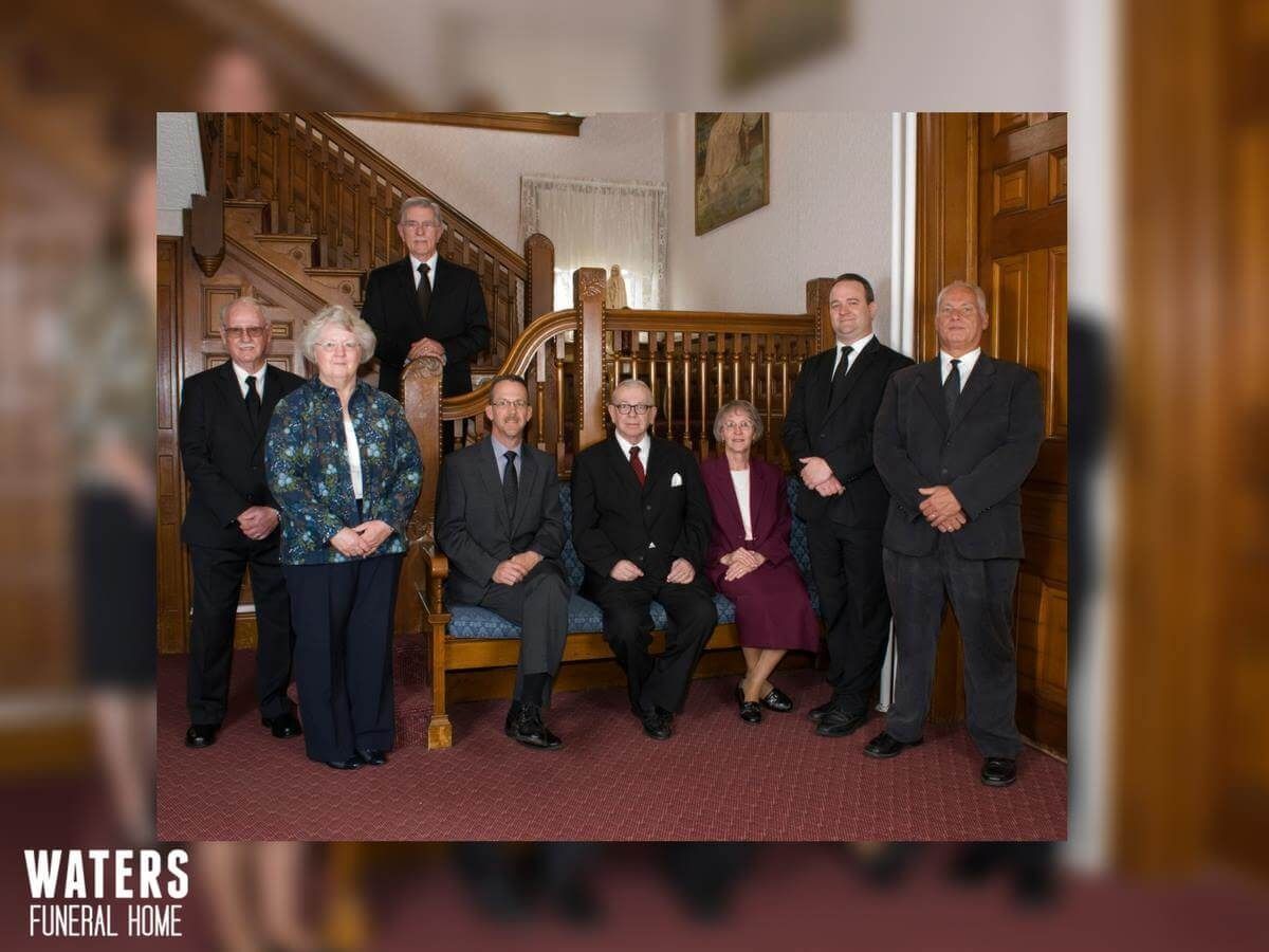 Formal group portrait of seven people in suits and dresses on a staircase and lobby, with a fireplace behind them
