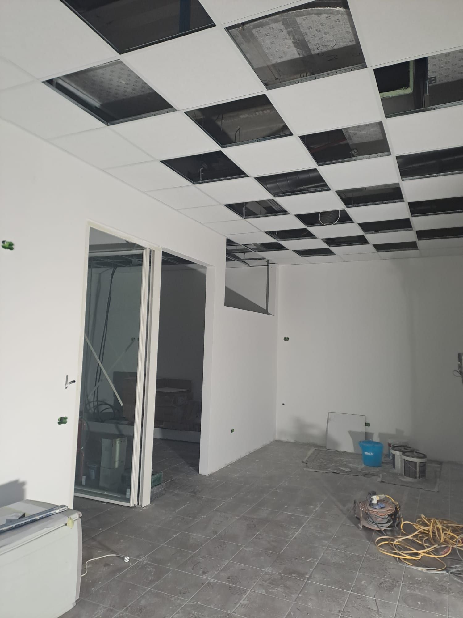 Stanza vuota e incompiuta, con controsoffitto a piastrelle a scacchi bianche e nere, porta aperta e materiali edili sul pavimento