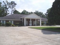 Single-story brick building with white-columned entrance and a large paved driveway, surrounded by trees