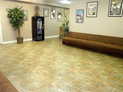 Empty lobby with beige tile floor, tan walls, potted plants, framed art, and a brown bench near a water cooler.