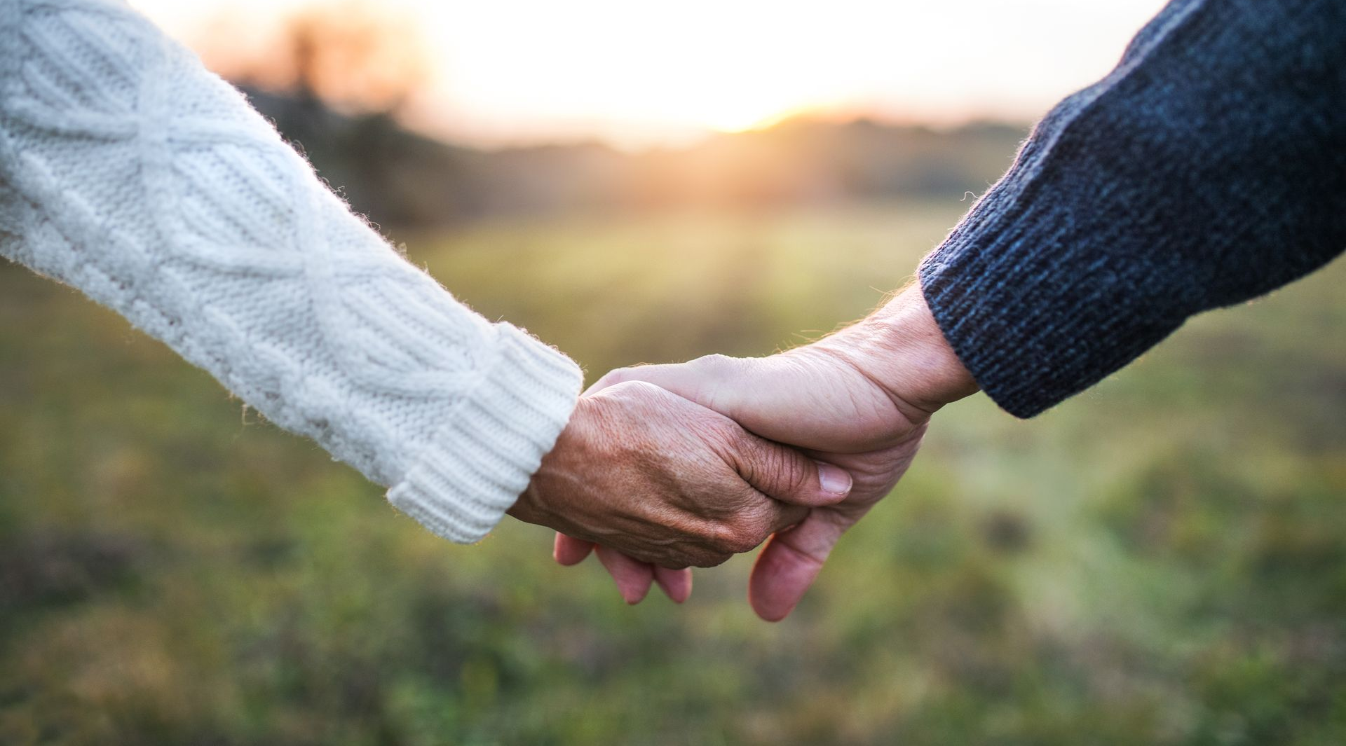 Two people holding hands outdoors, one in a white sweater and one in a dark sweater, at sunset.