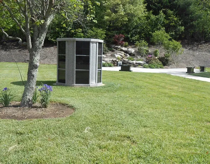 Small gray utility shed beside a tree on a green lawn with landscaping and a walkway in the background