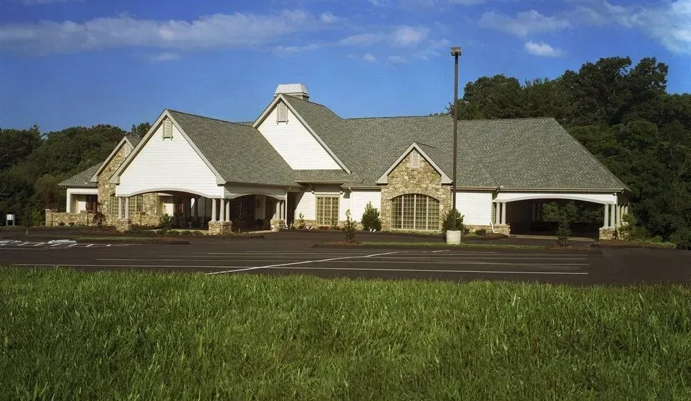 Large white house with gray roof and stone entry, seen from a grassy roadside under a blue sky.