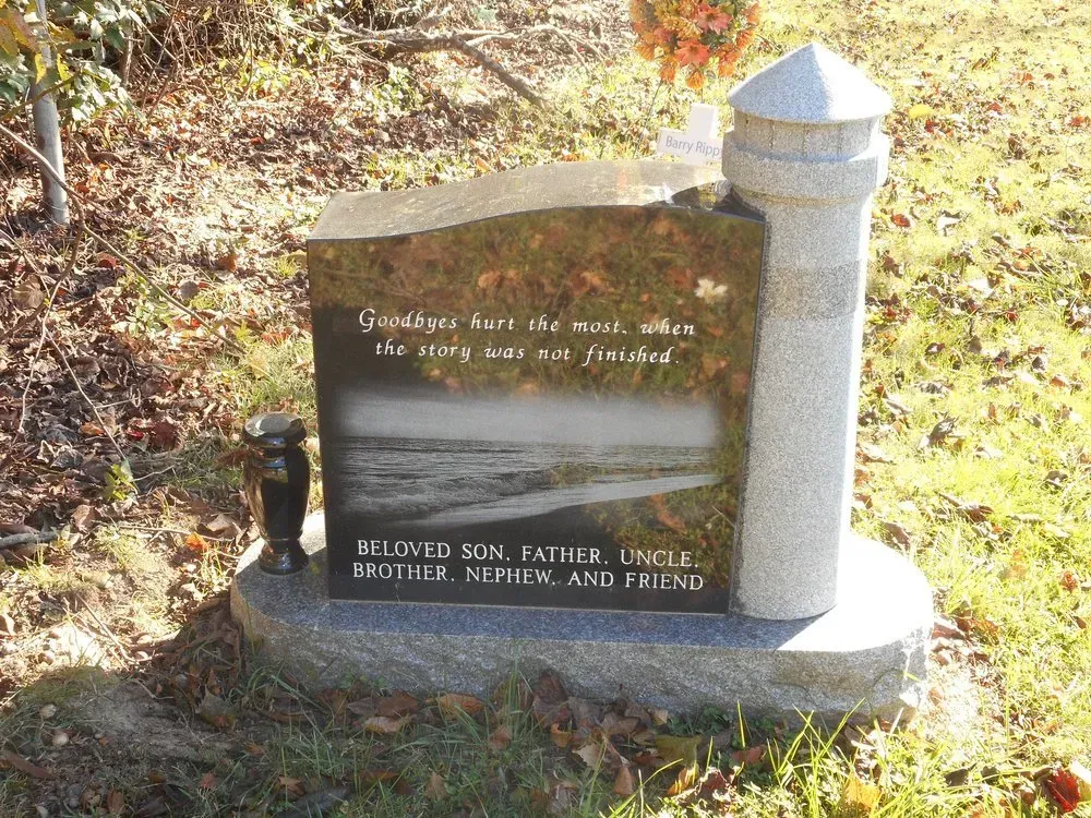 Memorial plaque and stone post by a wooded roadside, with a river scene etched on the plaque