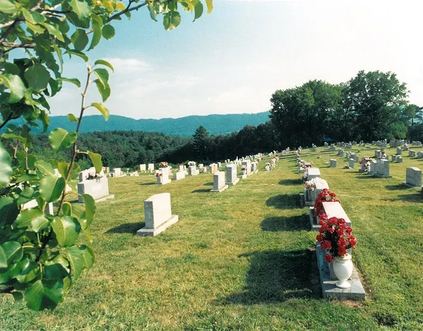 A cemetery with rows of white headstones on a grassy hill, bordered by trees and distant blue mountains.