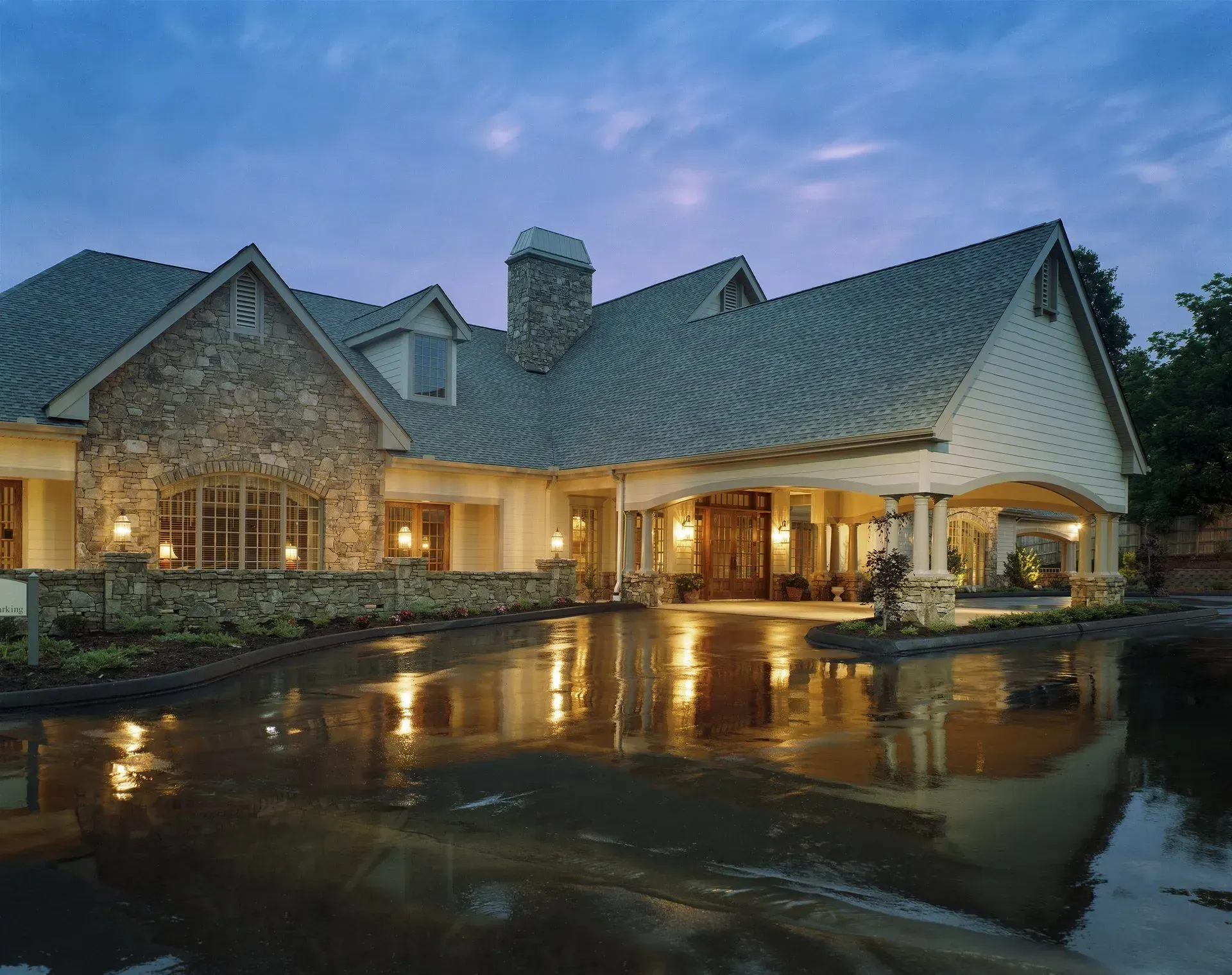 A warmly lit stone house with a steep roof reflected in wet pavement at dusk.