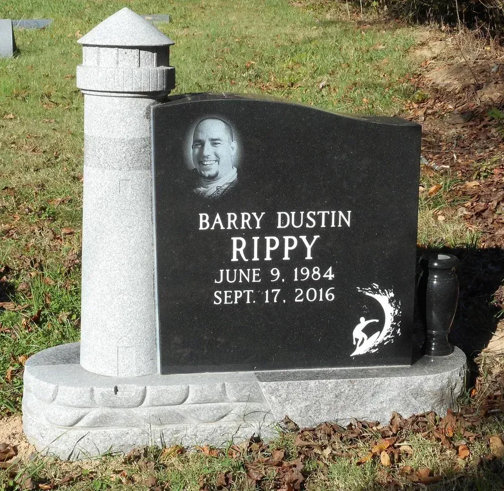 Black granite memorial headstone with a portrait, lighthouse design, and engraved name and dates on a grassy lawn