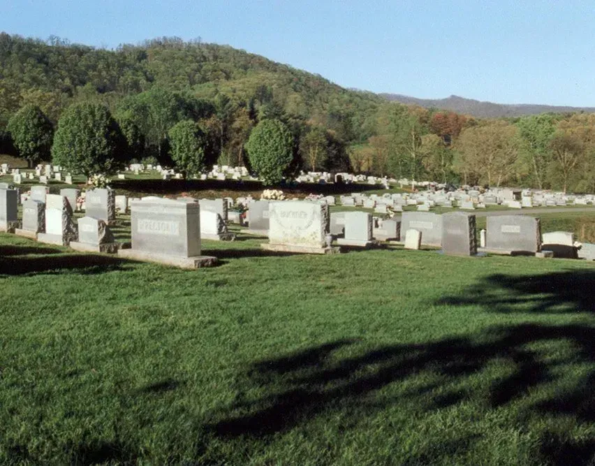 Rows of white gravestones in a grassy cemetery with wooded hills in the background.
