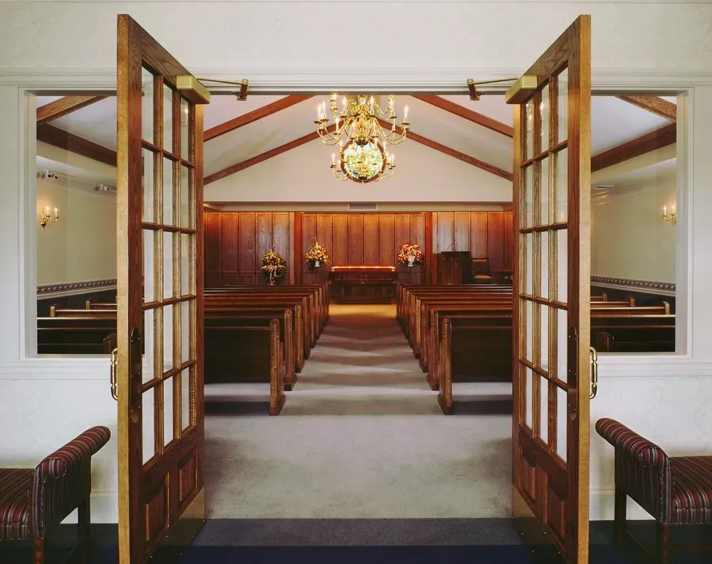 Open church doors leading to a pew-lined aisle and chandelier-lit altar in warm wood tones