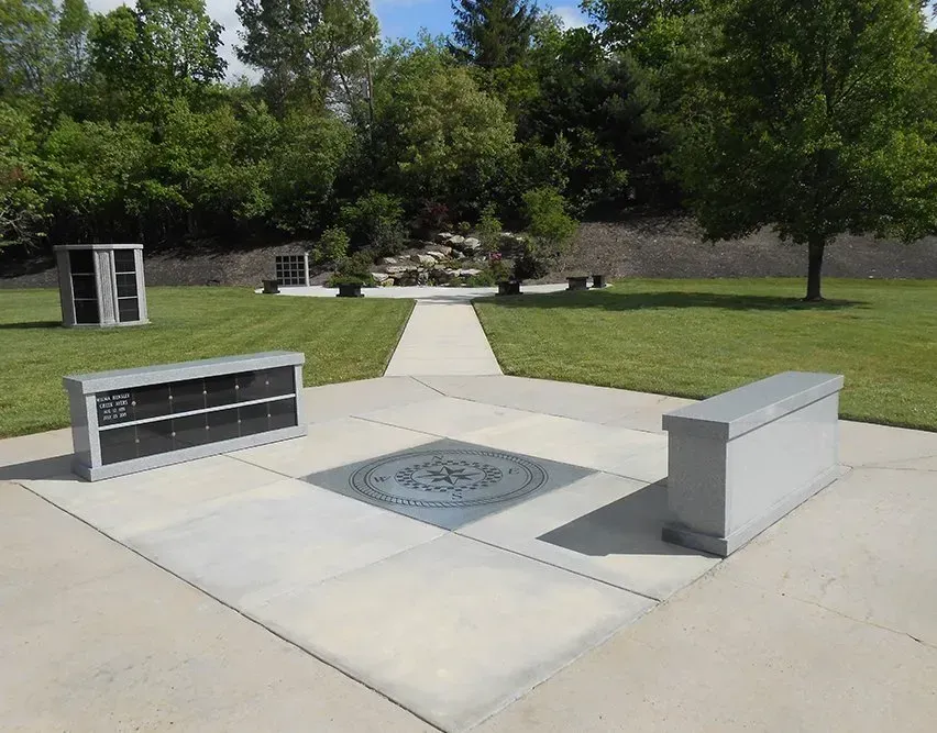 Outdoor memorial plaza with benches, engraved stone center, and a walkway amid grass and trees