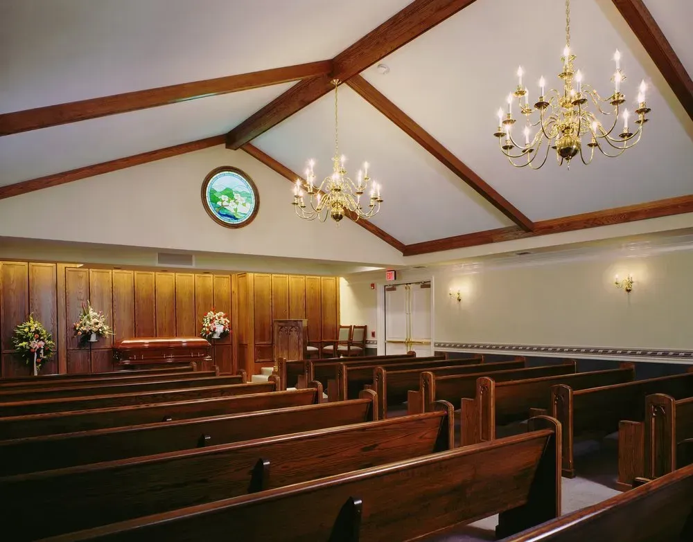 Empty church sanctuary with wooden pews, chandeliers, and a stained-glass window above the altar.