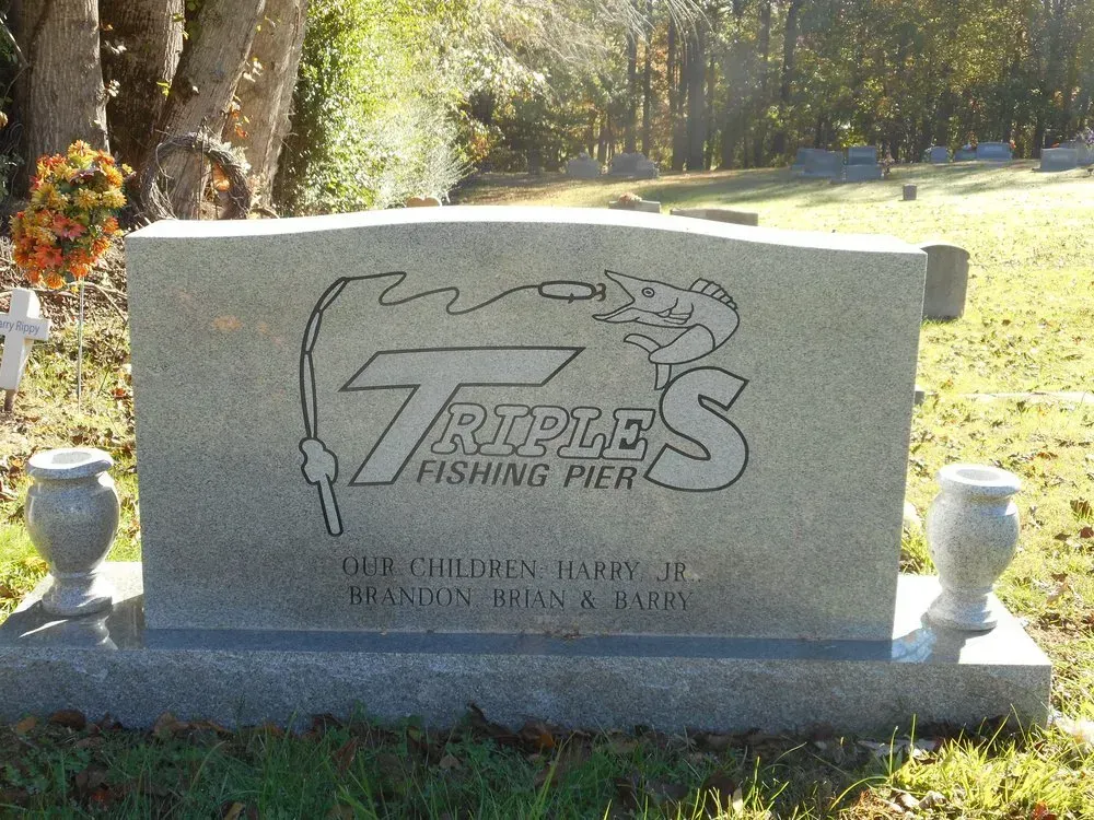 Gray granite grave marker engraved “Trout’s Fishing Pier,” with fishing pole graphic in a sunny cemetery.