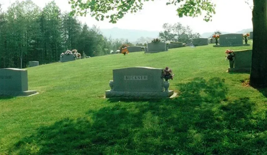 Grassy cemetery hillside with headstones and flower arrangements under trees on a sunny day