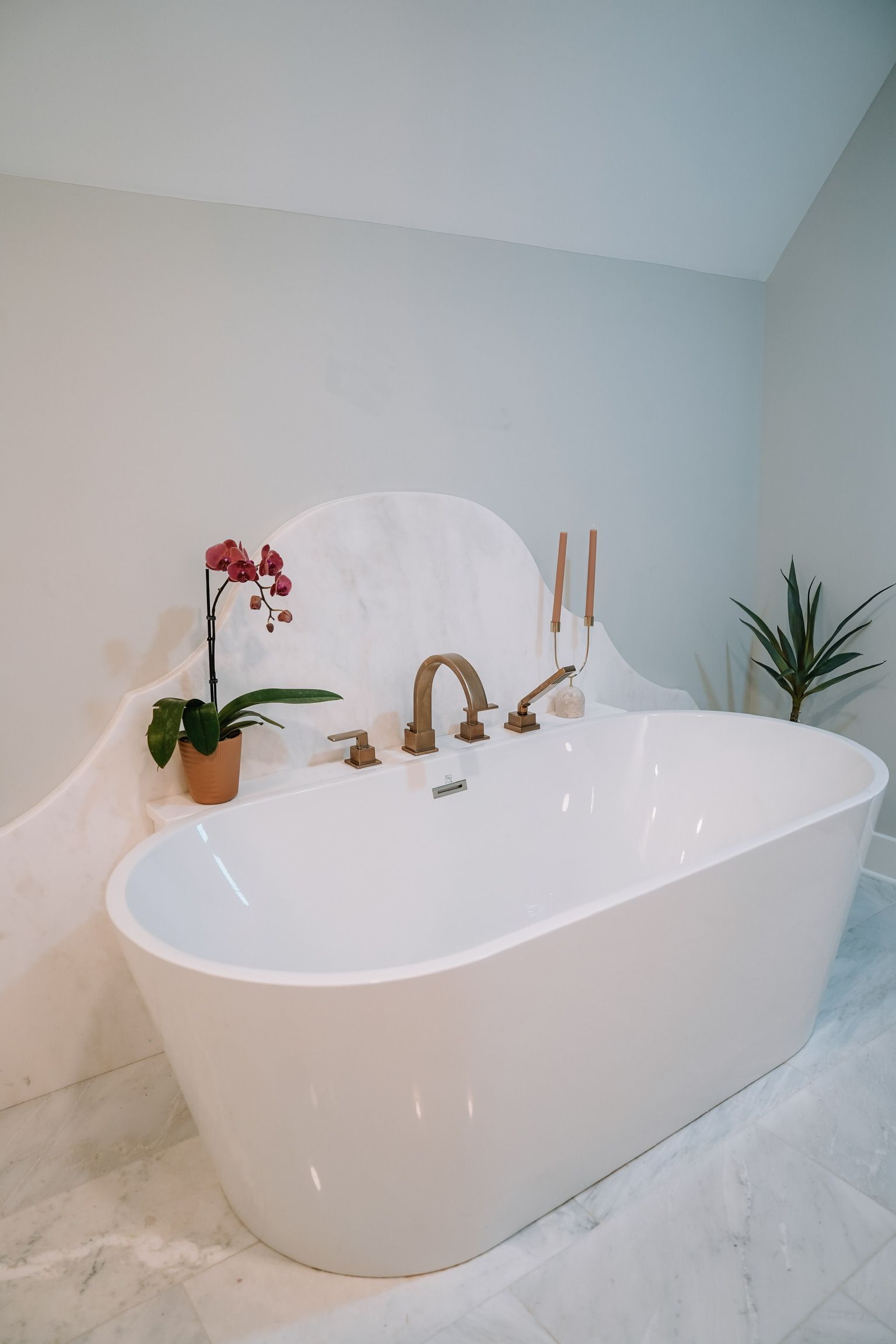 A white freestanding bathtub set against a curved marble accent wall with gold fixtures, an orchid, and a small plant.