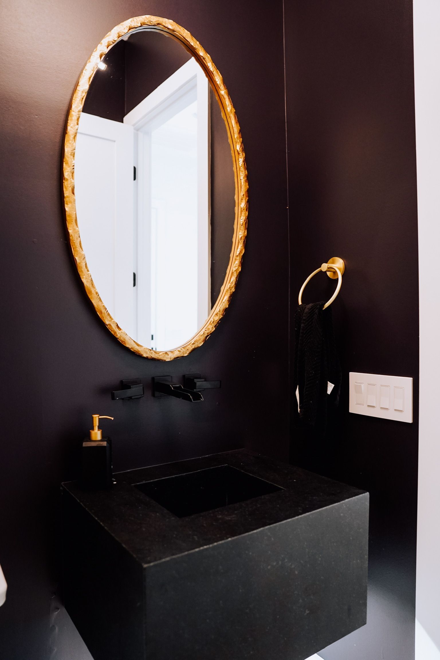 A modern bathroom features a dark wall, a gold oval mirror, a black floating sink, and a matching gold towel ring.