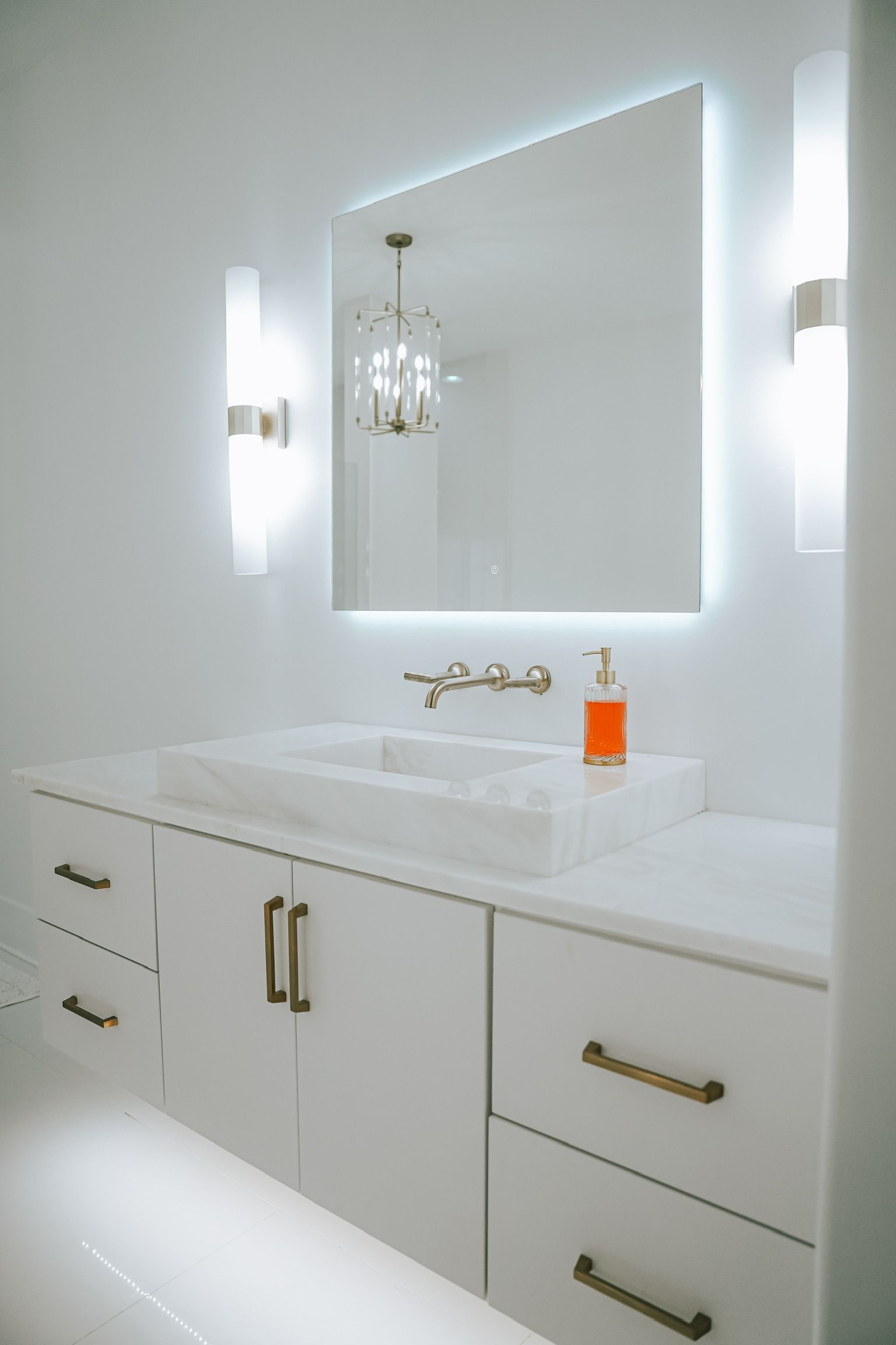A modern white bathroom vanity with gold hardware, a rectangular back-lit mirror, and sconce lighting on a white wall.