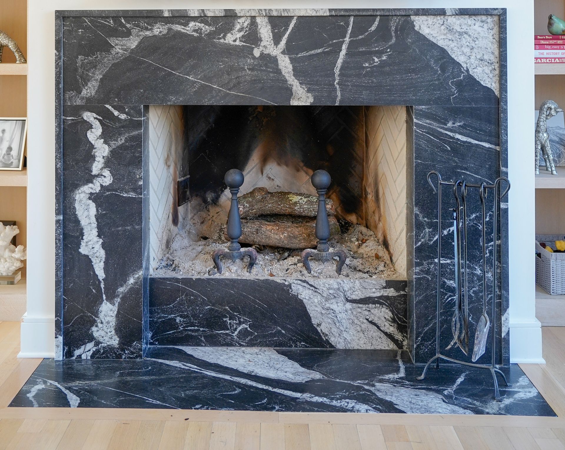 A black marble fireplace with white veining, featuring iron andirons and a tool set, set against white walls.