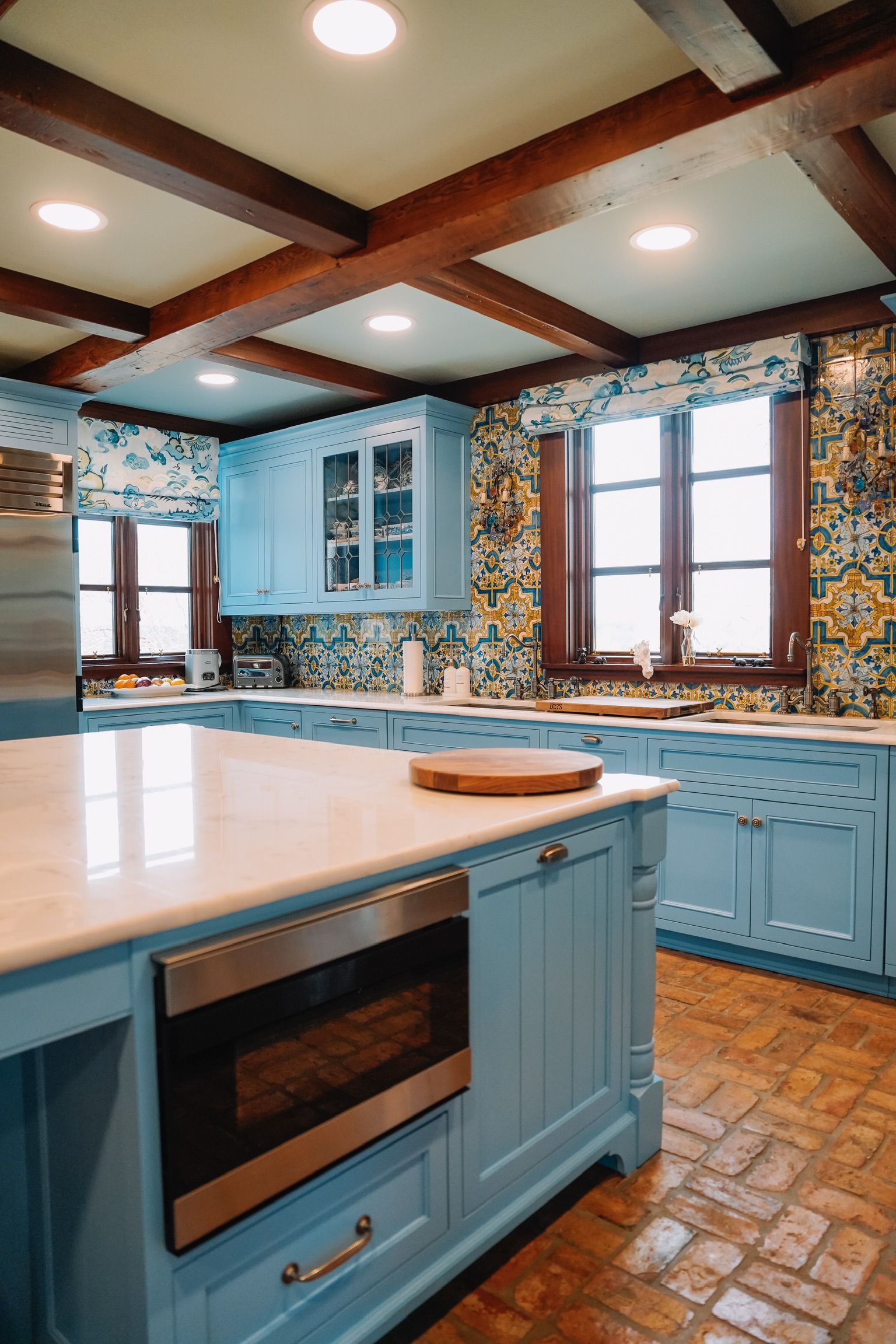 A rustic kitchen features blue cabinetry, an island with a built-in microwave, tiled backsplashes, and brick flooring.