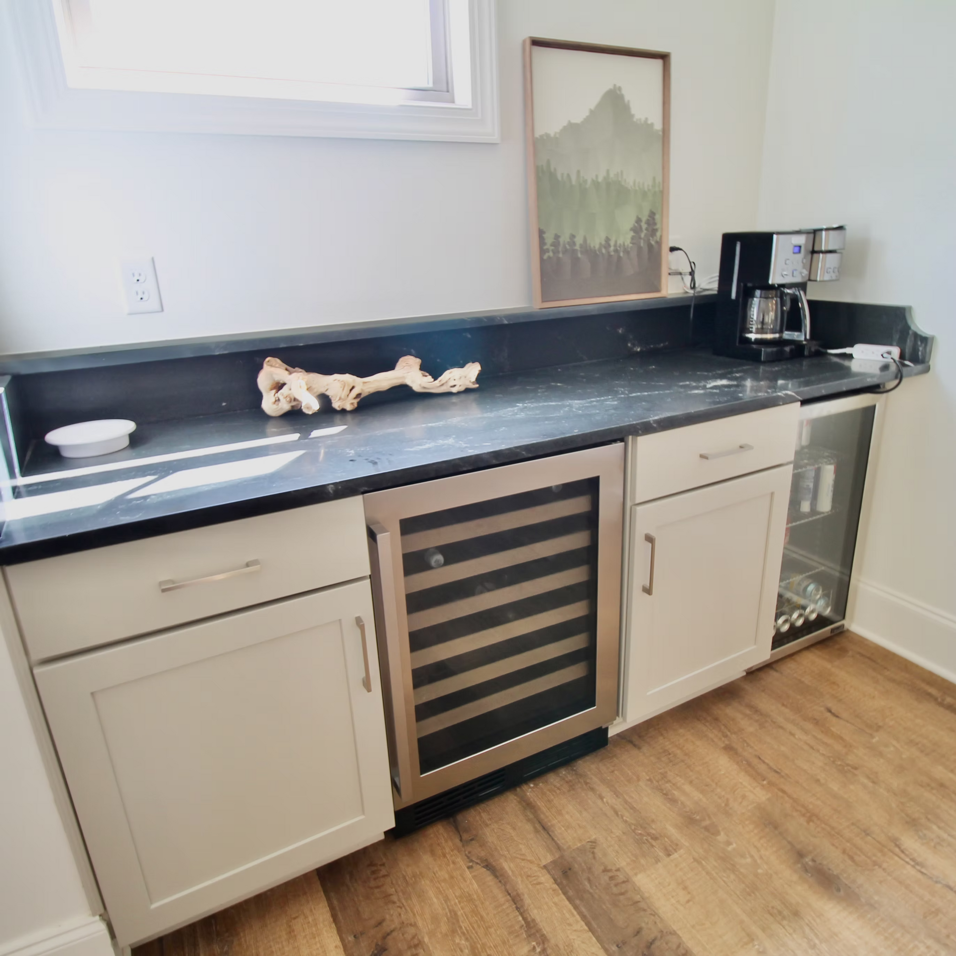 White cabinets with wine fridge and coffee maker on a black countertop; driftwood decor, framed art, and a small window.
