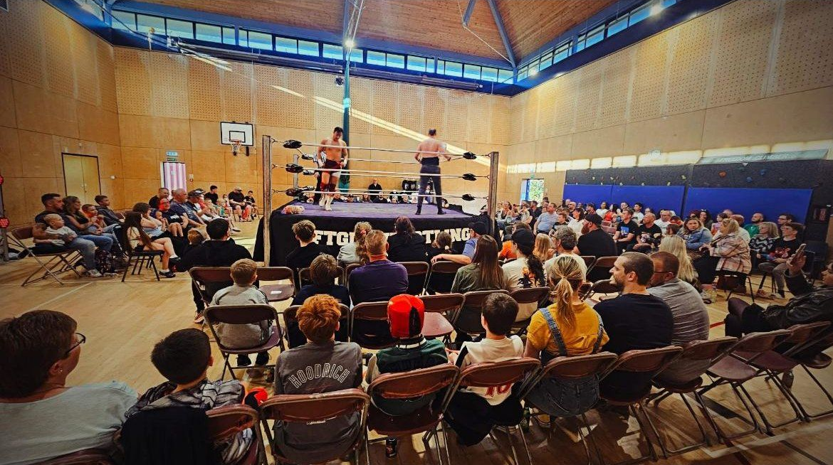 Wrestling match in a gymnasium, with a ring centered and spectators seated around it.