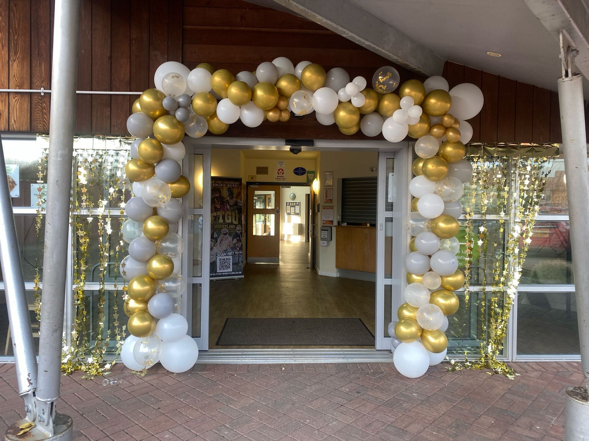 White, gold, and silver balloon arch framing a building entrance.
