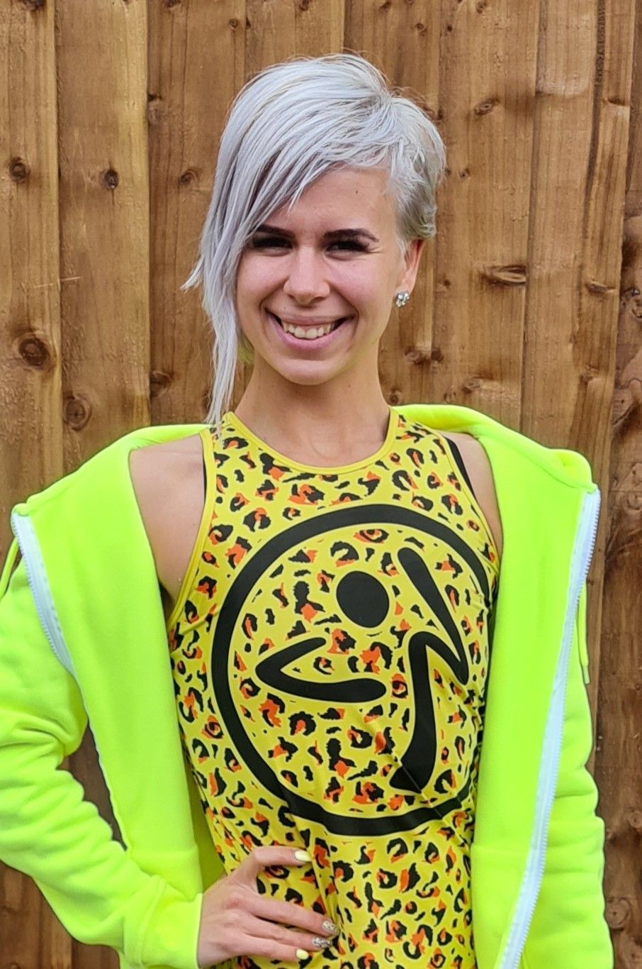 Person smiling in a neon yellow outfit with silver hair, standing against a wooden fence.