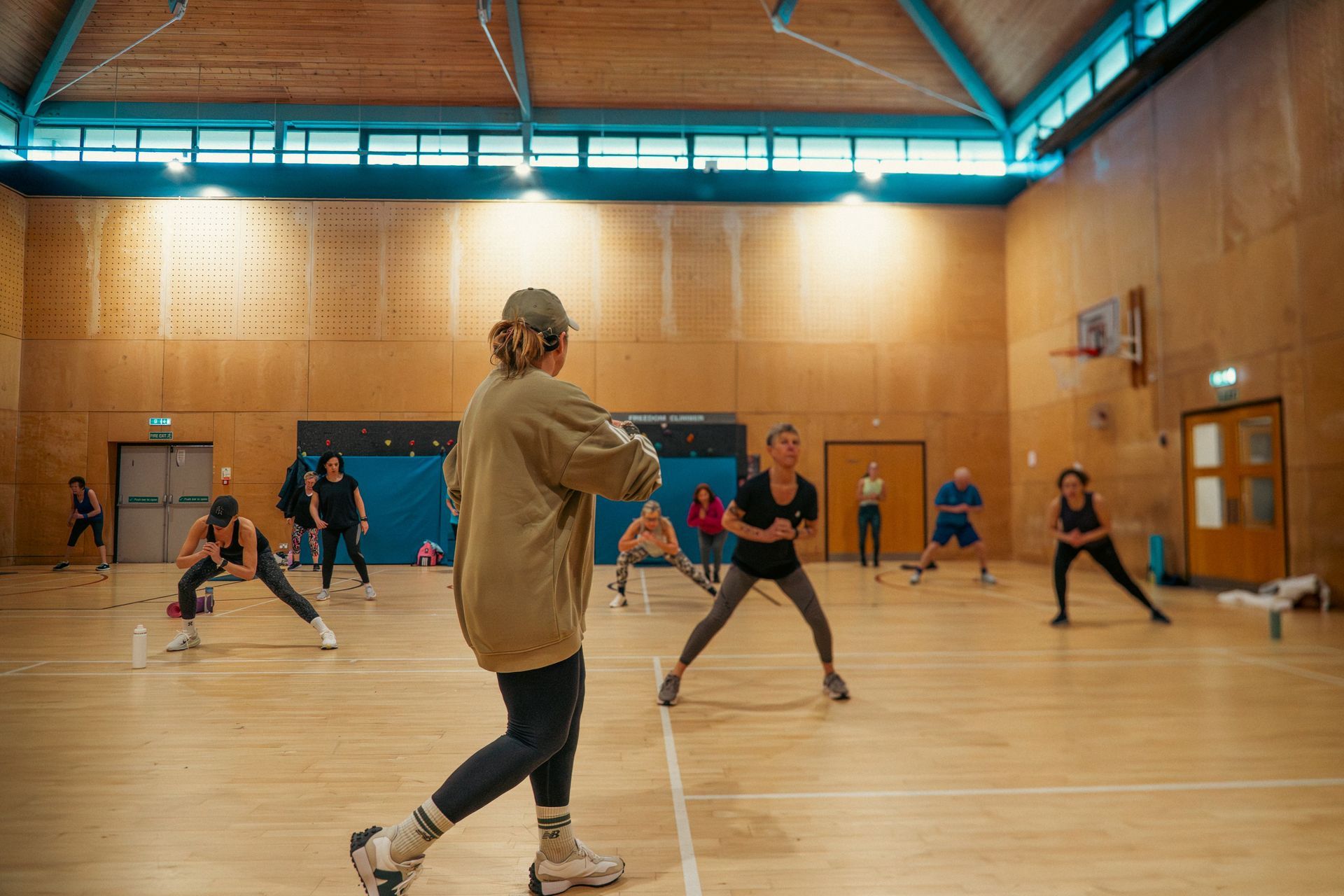 Indoor gym class with people stretching on a wooden court under bright skylights