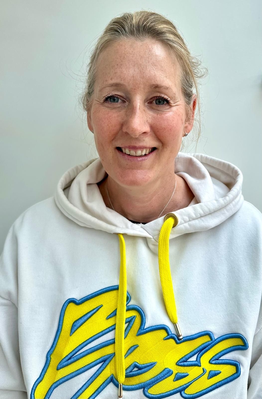 Smiling person in a white hoodie with yellow drawstrings and blue-yellow lettering, against a light background