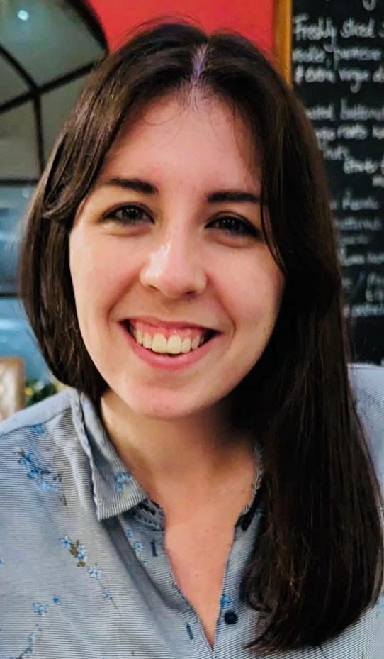 Smiling woman with short dark hair in a blue shirt, standing indoors near a chalkboard menu.