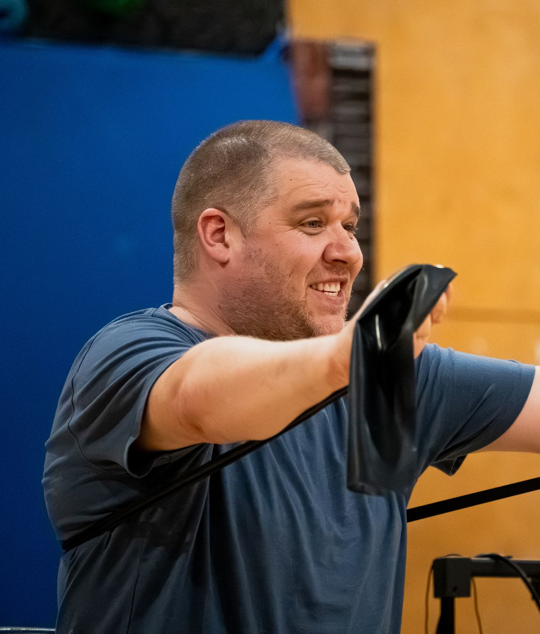 Man in a blue shirt smiling while lifting black straps in a gymnasium