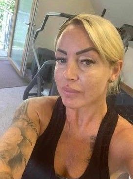 Woman taking a selfie in a gym, wearing a black tank top with workout equipment in the background