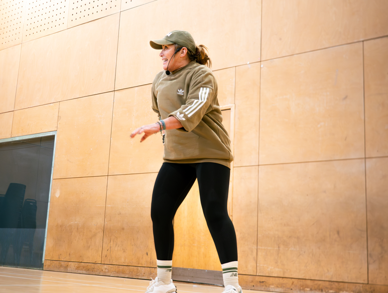 Person in a tan sweatshirt and black leggings standing in a gym, mid-gesture during a workout