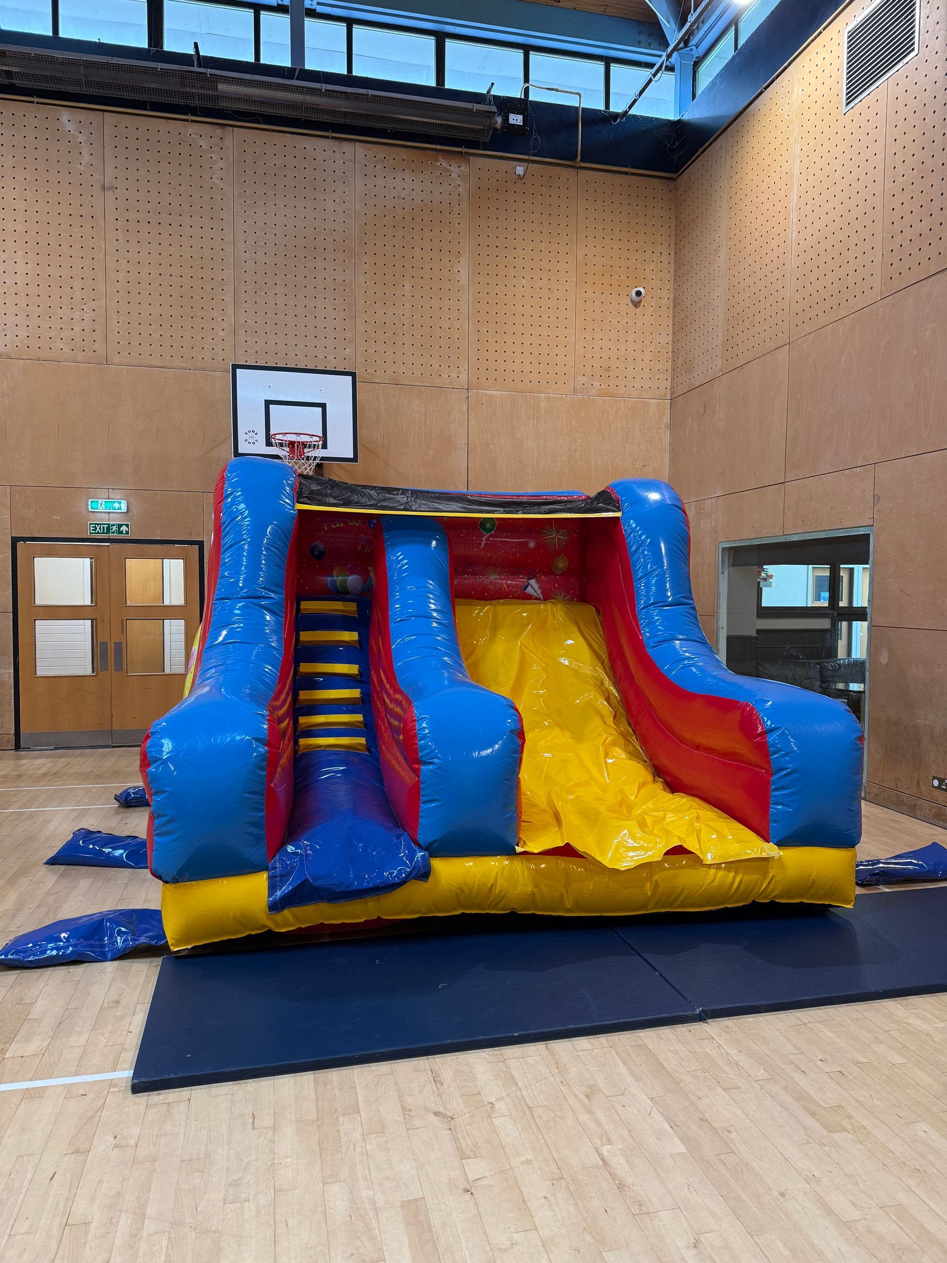Blue and yellow inflatable slide in a gymnasium with a basketball hoop and wooden walls