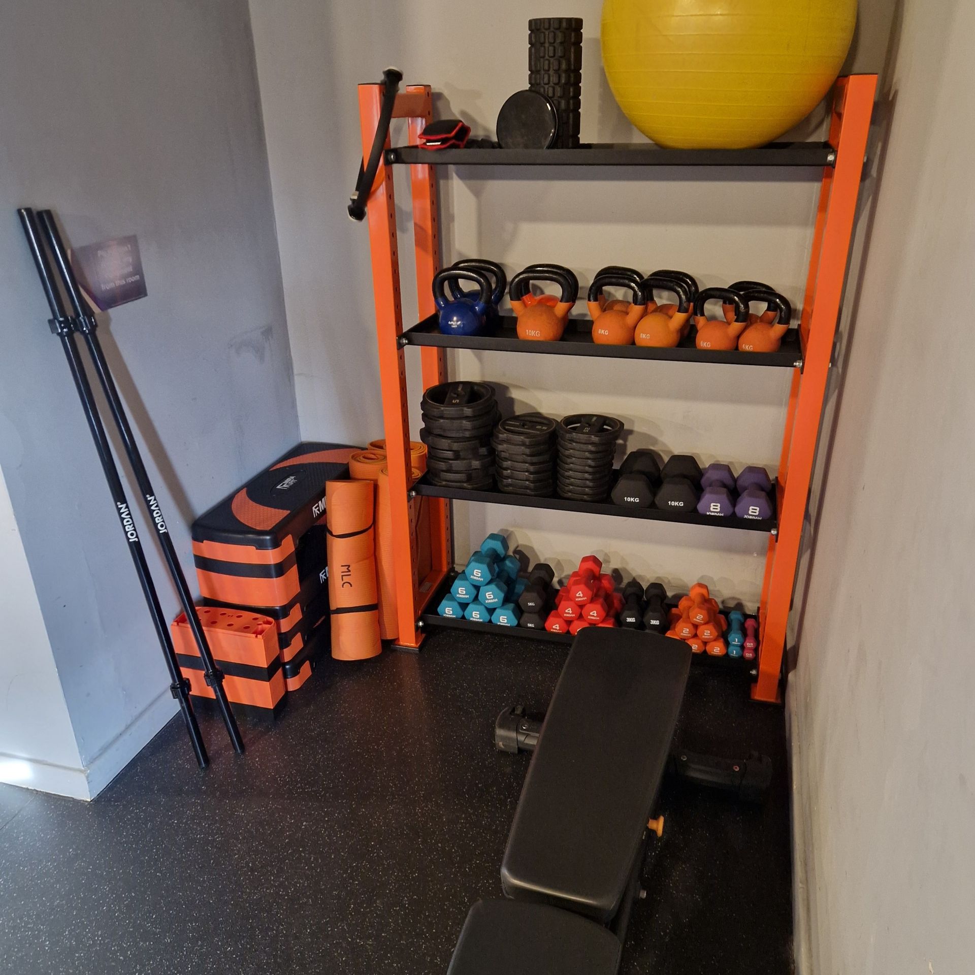 Home gym corner with kettlebells, weights, foam rollers, and exercise equipment on orange shelving.