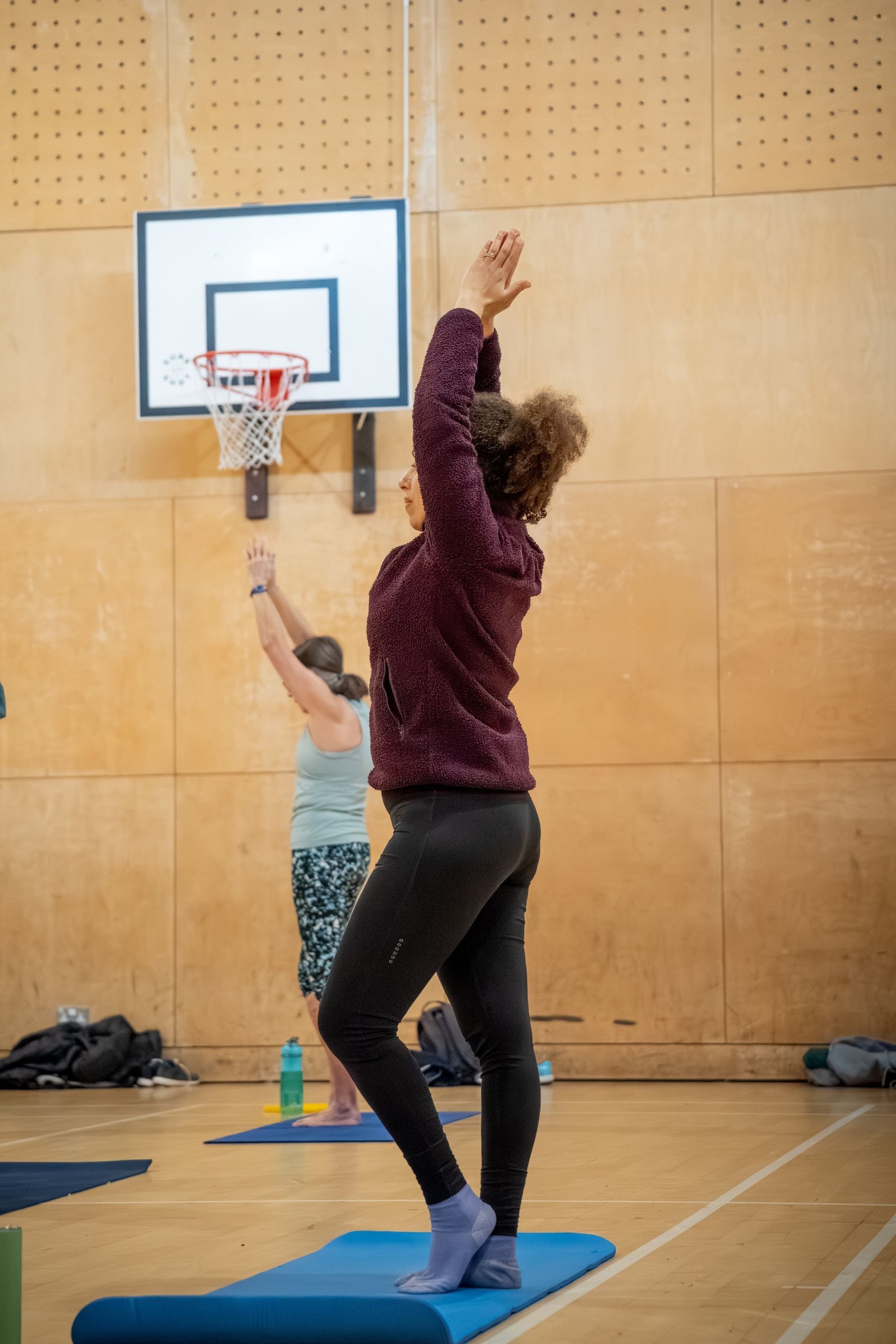 Two people doing yoga poses on mats in a gym, one balancing with arms raised near a basketball hoop.