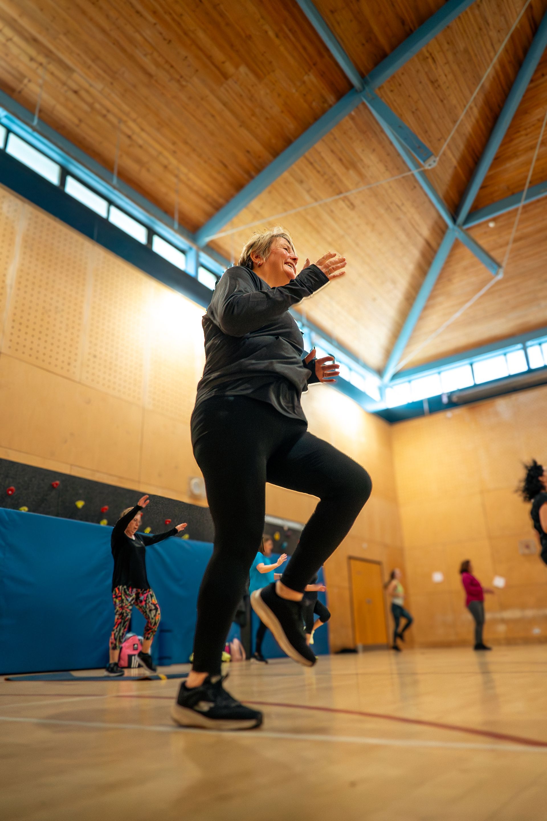 Person dancing in a gymnasium with a wooden ceiling and other people in the background