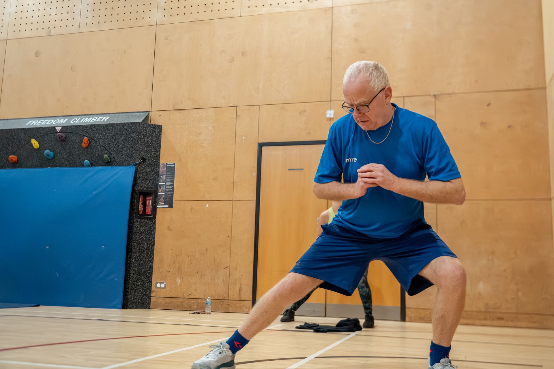 Man in blue shirt and shorts practicing a side lunge on a gym floor, holding a small ball