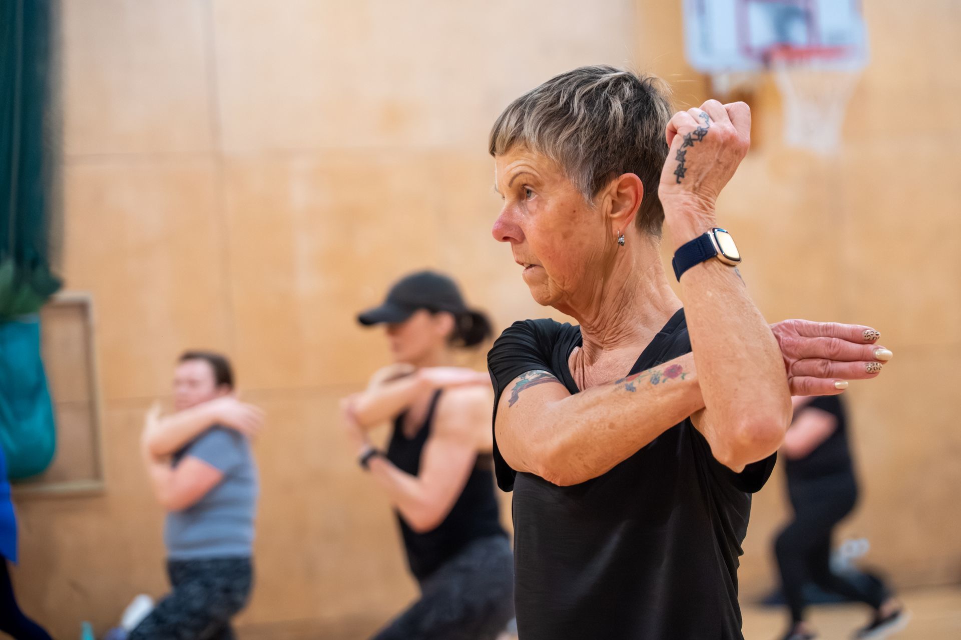 People exercising in a studio, with a woman in black stretching her arm in the foreground.