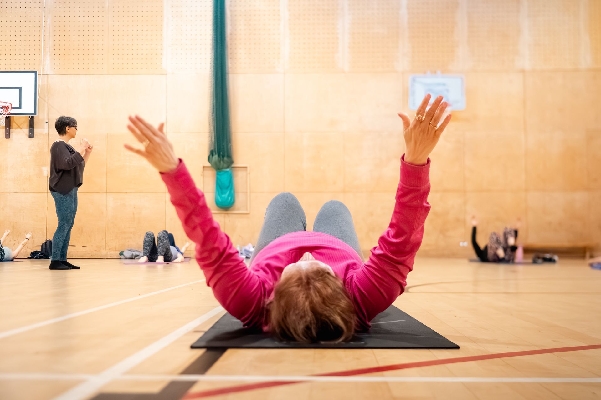 Person lying on a gym mat with arms raised in a gymnasium, another person standing nearby