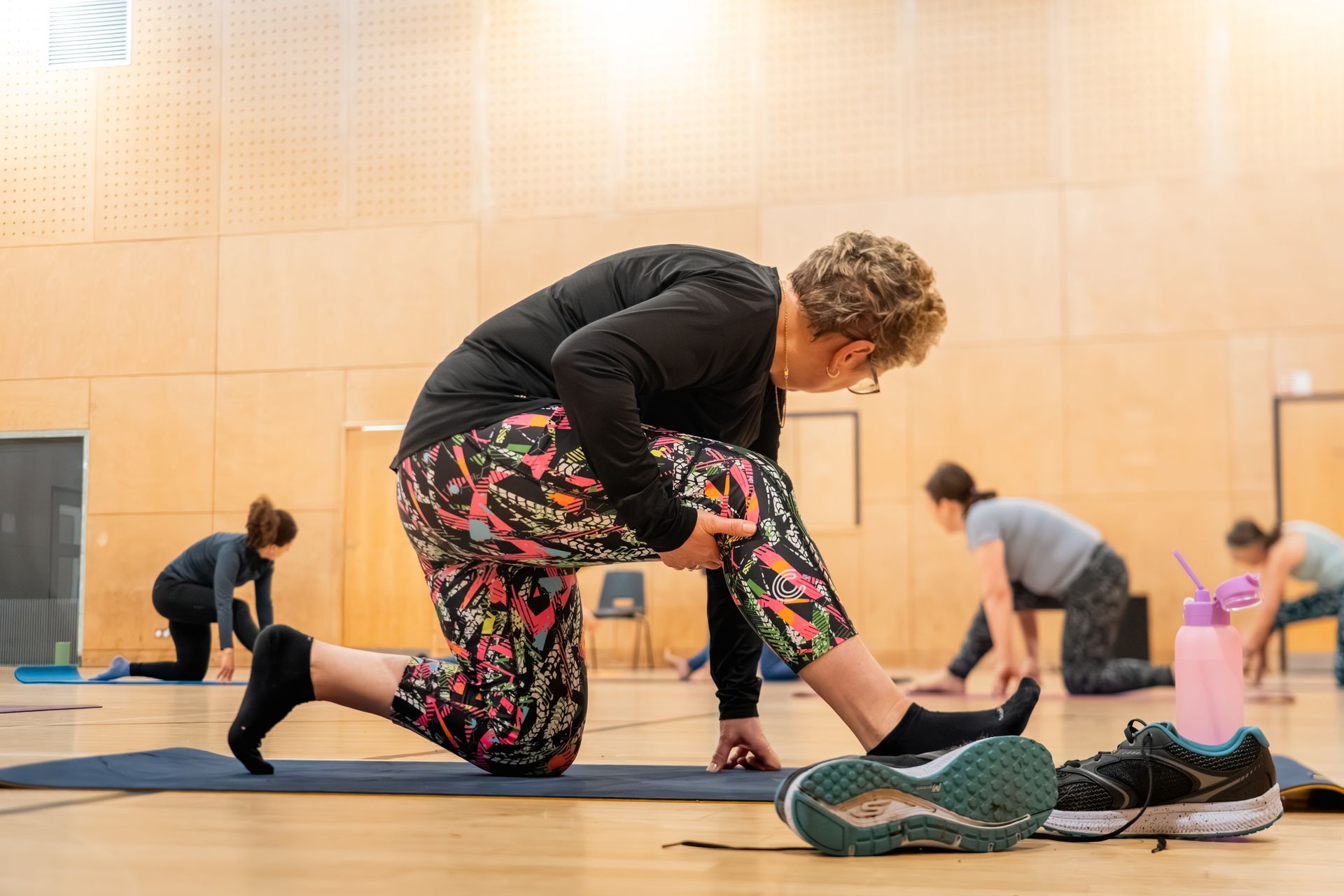 People stretching on mats in a gym, with one person in the foreground adjusting a knee stretch.