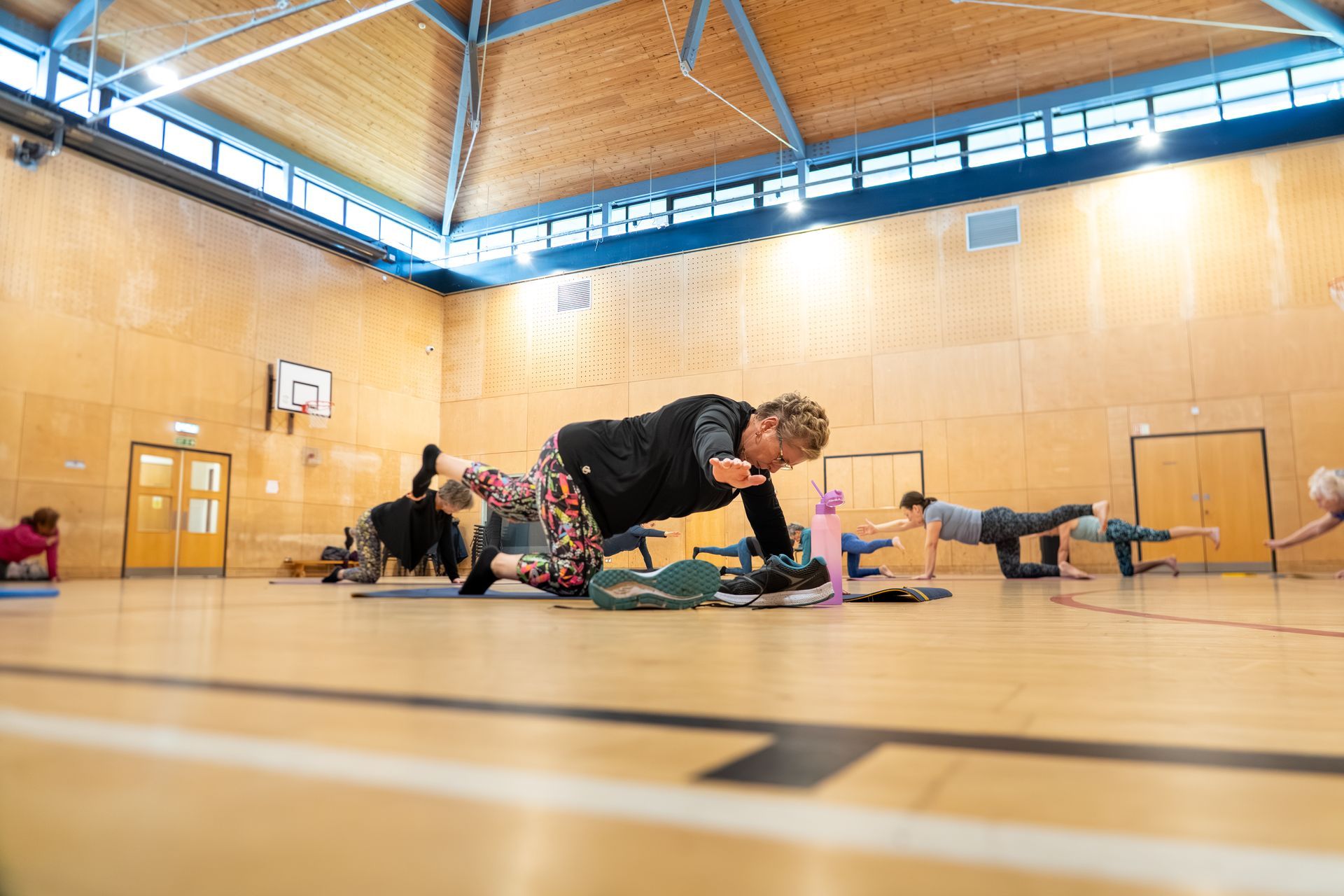 People doing yoga poses in a bright gymnasium with wooden walls and a blue ceiling