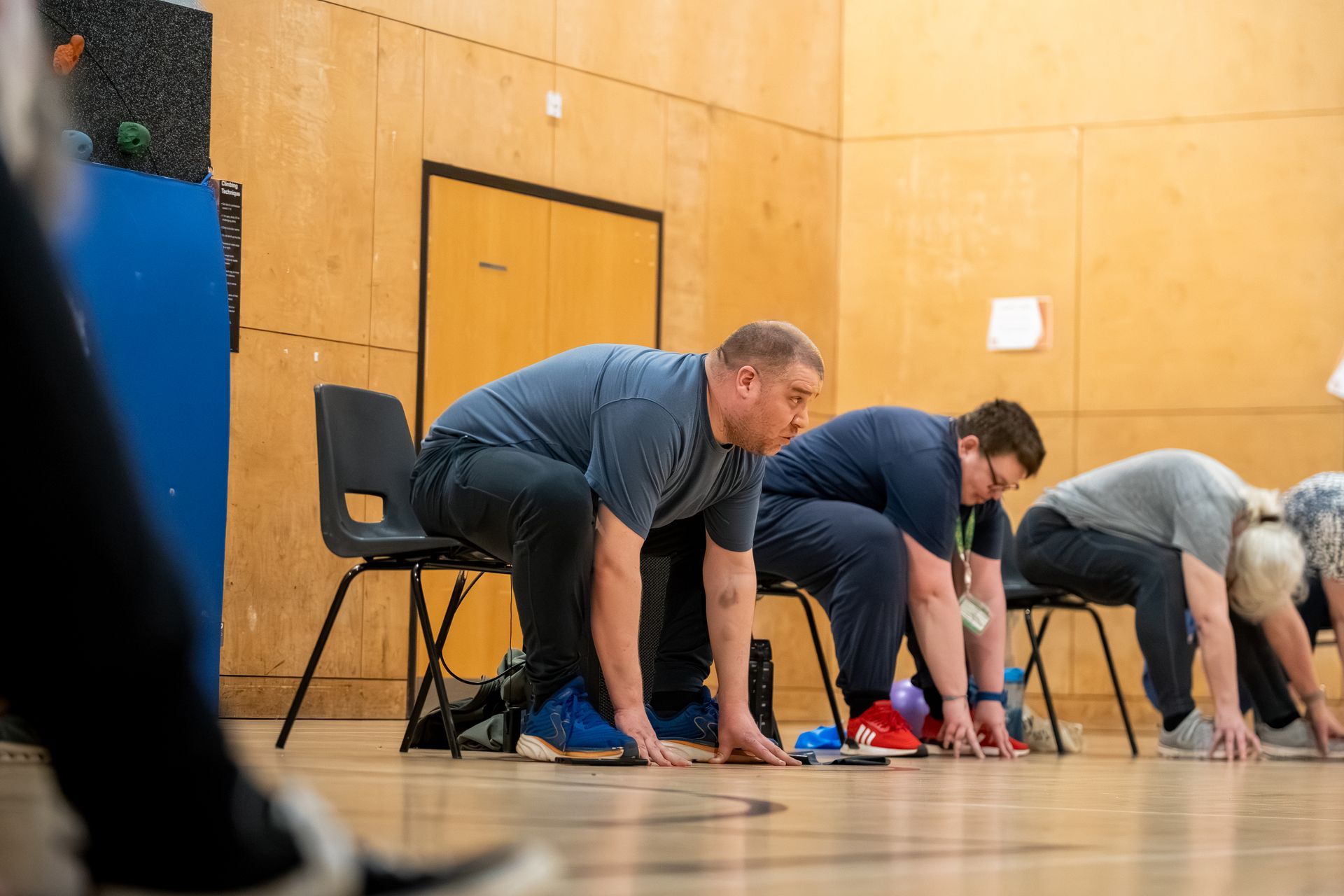 People stretching on a gym floor beside chairs and a wooden wall