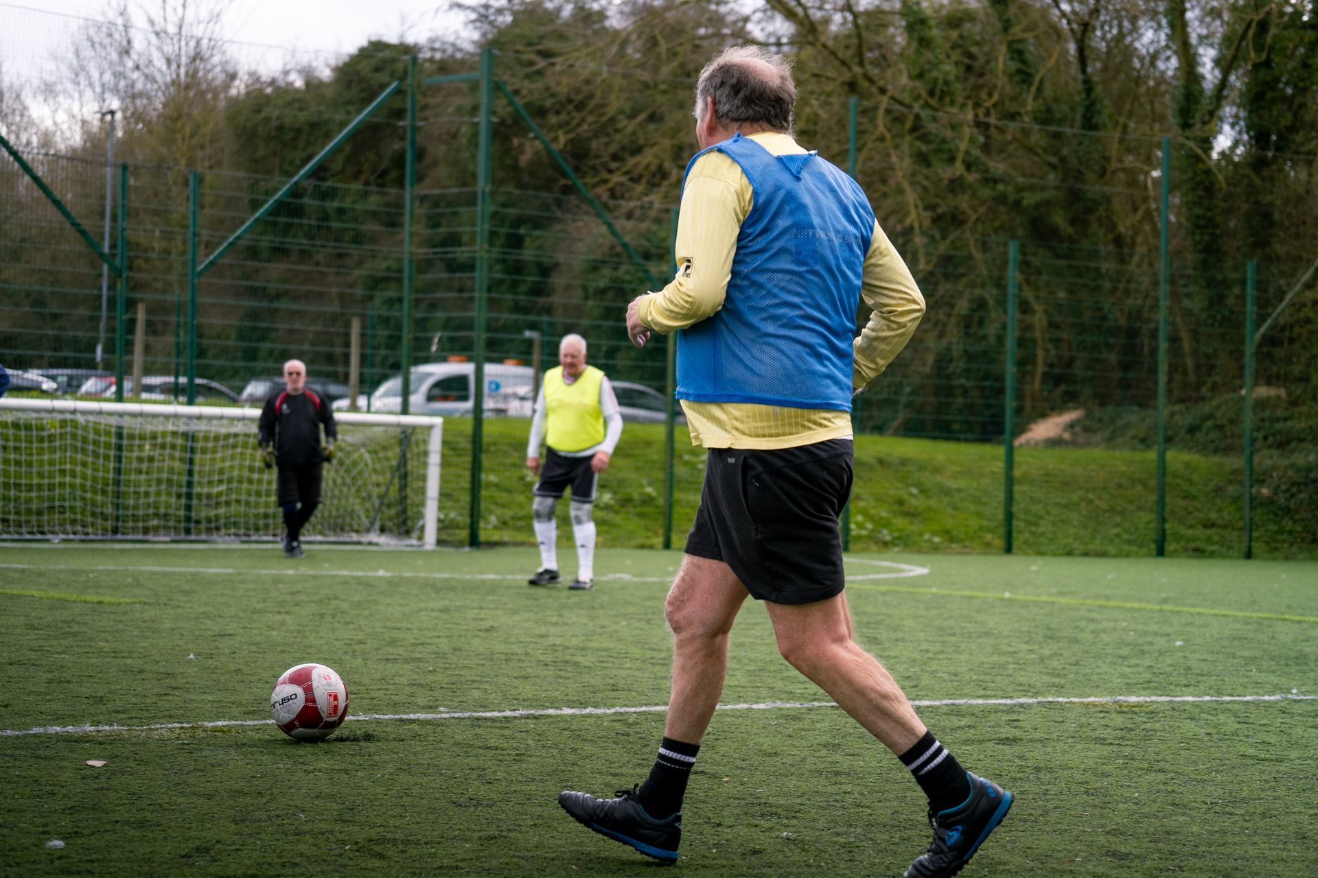 Soccer players on a field, with one in a blue vest approaching a ball near the goal